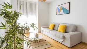 Ein modernes Wohnzimmer mit einem grauen Sofa, zwei gelben Kissen, einem Glastisch, einer gewobenen Stehlampe und einer großen Pflanze.