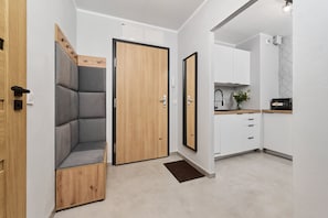 Un intérieur moderne avec une porte en bois, un banc rembourré de couleur grise, un coin cuisine doté de meubles blancs et un sol carrelé.