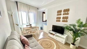 Een moderne woonkamer met een grijze bank, een flatscreen televisie, een eethoek met een tafel en stoelen, en een potplant.