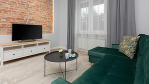 Un salón con pared de ladrillo, un televisor de pantalla plana sobre un mueble blanco, un sofá verde y una mesa de centro redonda.
