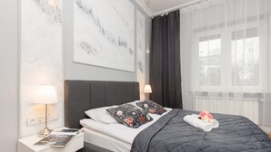 Un dormitorio ordenado con cama, almohadas, una mesita de noche con lámpara y una ventana con cortinas.