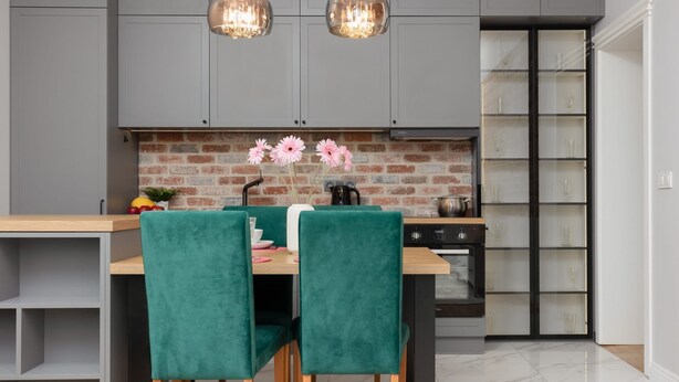 Una cocina moderna con armarios grises, salpicadero de ladrillo y un comedor con mesa de madera y sillas tapizadas en verde.