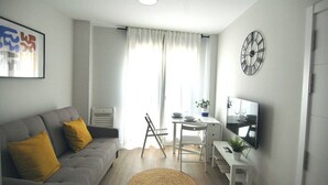 A modern living room with a grey sofa, a small white table, a TV, and a large window with curtains. - Vivodomo | New, great location, optional parking (Madrid)