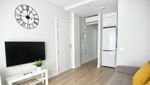 A modern living room with a flat-screen TV, a clock, a sofa, and a kitchen area with a refrigerator. - Vivodomo | New, great location, optional parking (Madrid)