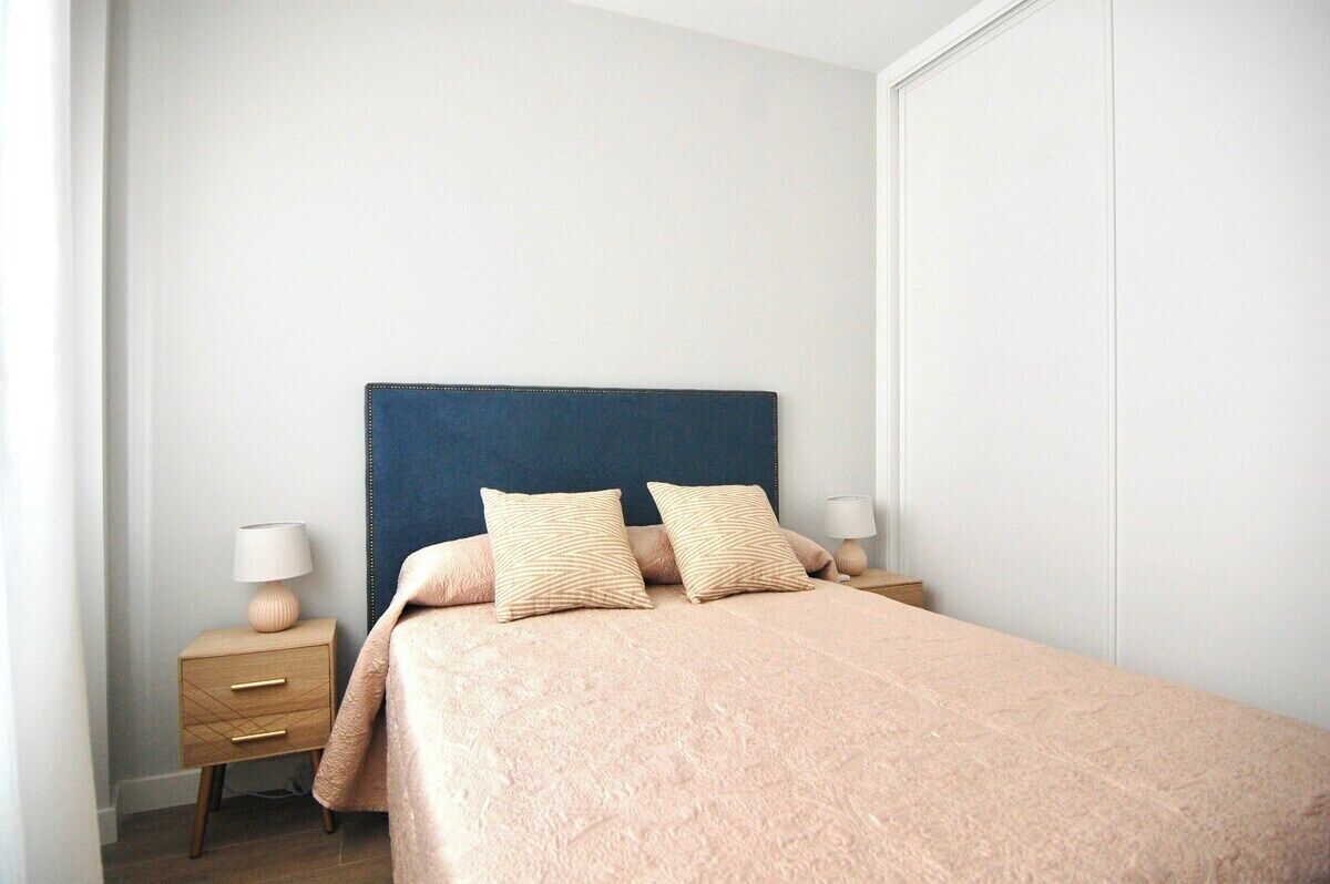 Ein ordentlich bezogenes Bett mit blauer Kopfteil und zwei Kissen, ein Nachttisch mit Lampe sowie ein Kleiderschrank in einem schlichten, sauberen Zimmer.