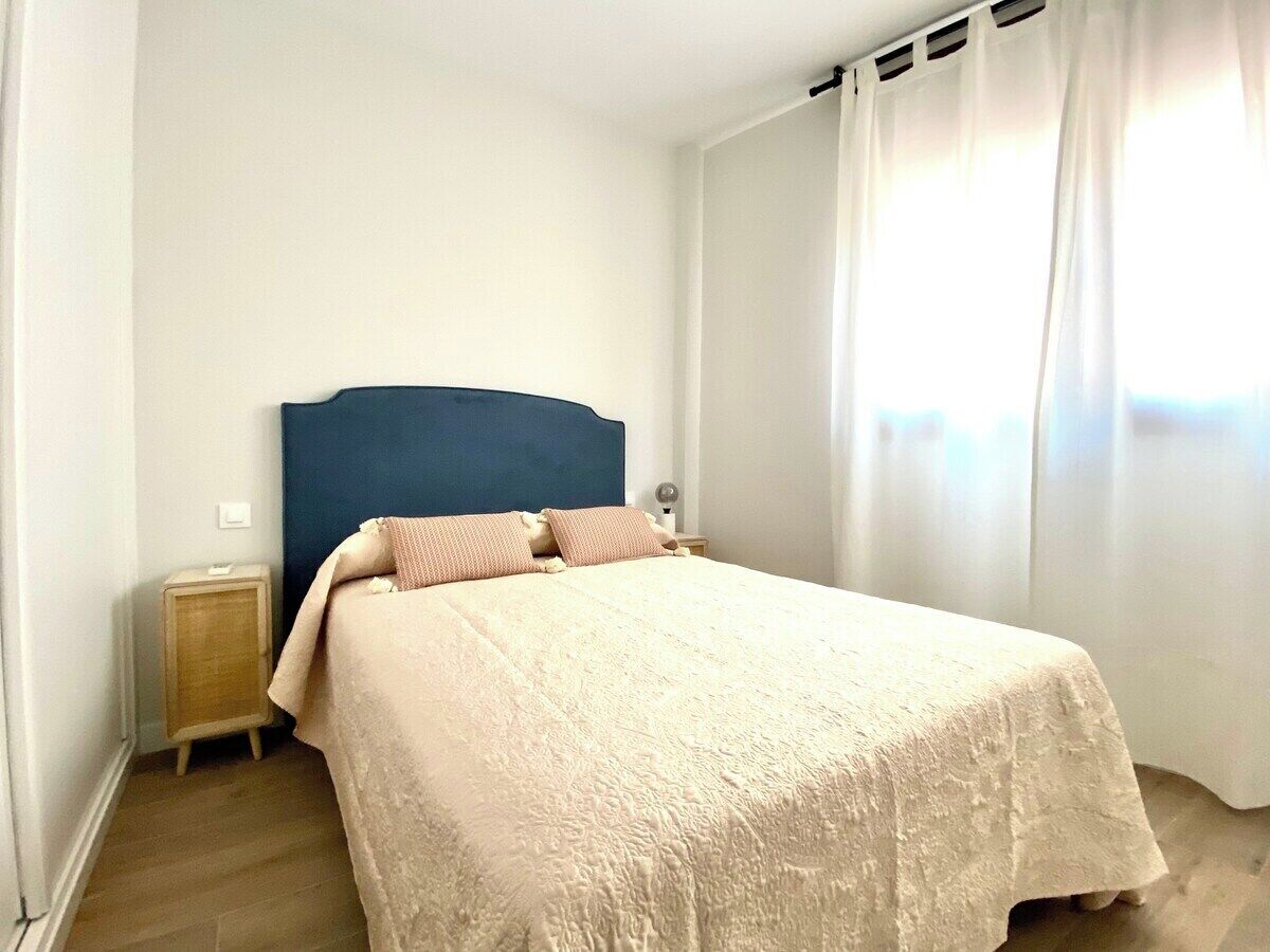 A bedroom with a bed, a nightstand, a blue headboard, and a window with curtains.