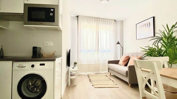 A compact living space with a washing machine, a microwave, a sofa, a dining table, and a potted plant. - Vivodomo | New, super location, optional parking (Madrid)
