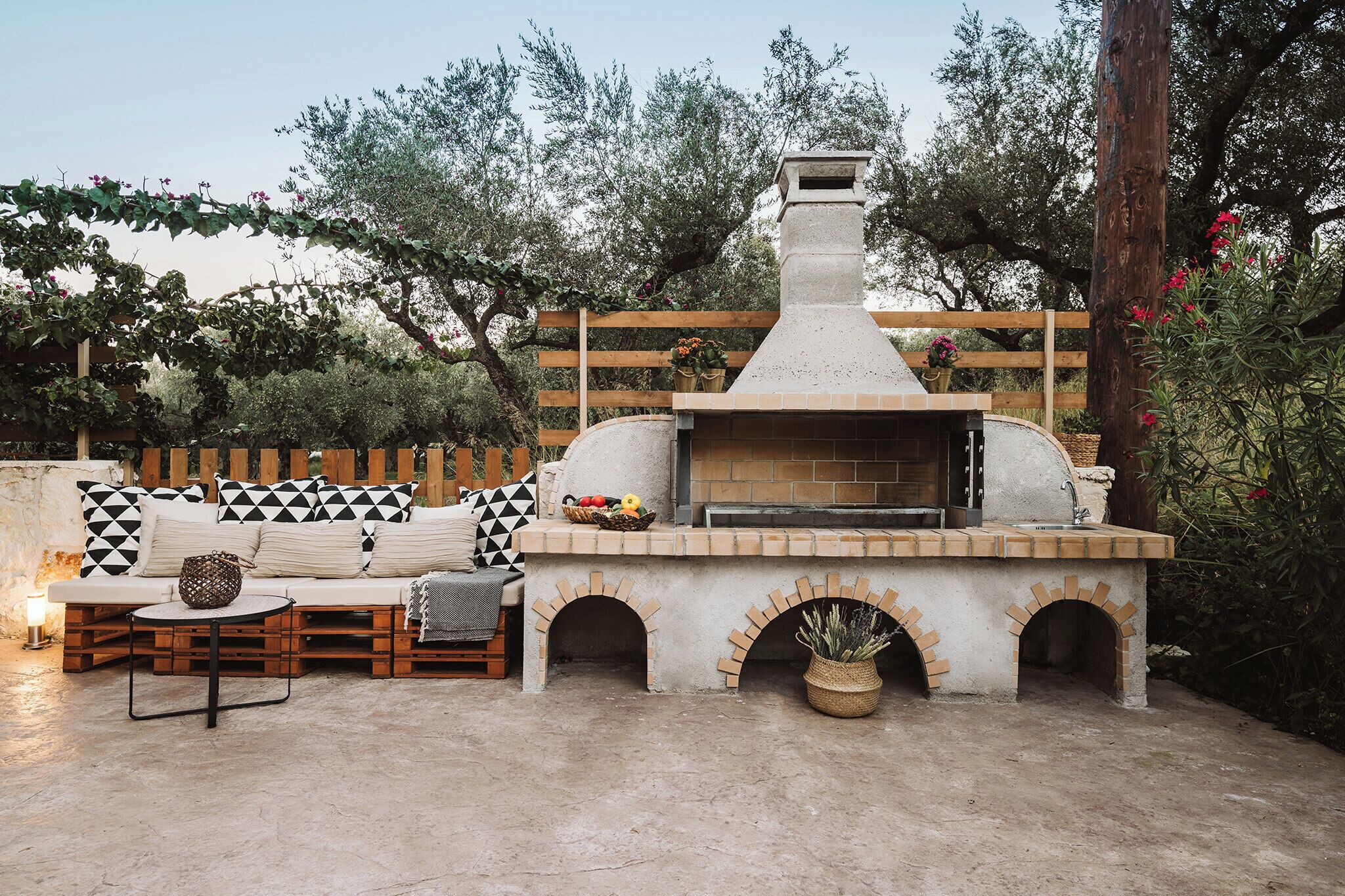 Een terras buiten met een ingebouwde pizzaoven, een houten bank met gestreepte kussens en een rustieke afrastering.