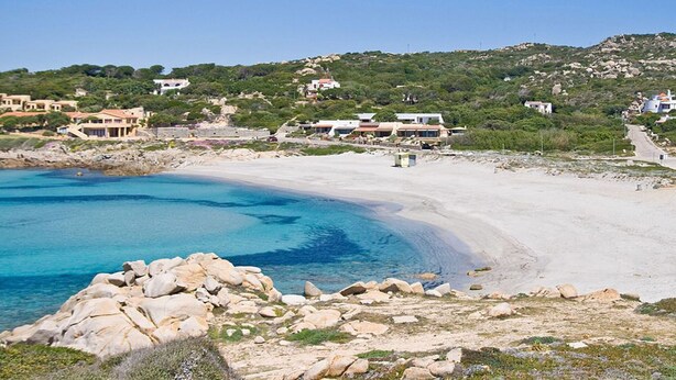 Una zona costera con una playa arenosa, costa rocosa y edificios al fondo.