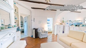Amplia sala de estar con un sofá, una silla y un banco. Hay una cocina con un refrigerador y un lavabo. La habitación tiene paredes blancas y piso de baldosas.