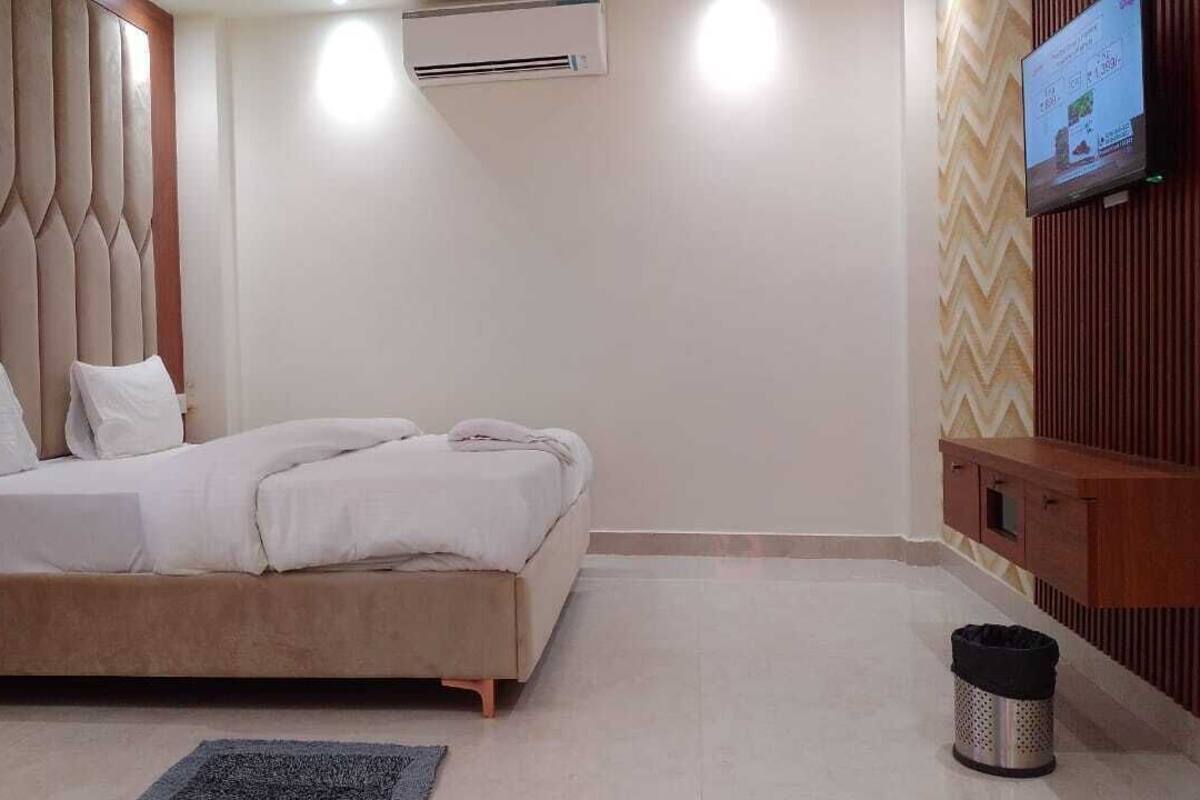 A bedroom with a bed, a TV mounted on the wall, an air conditioning unit, and a trash bin.