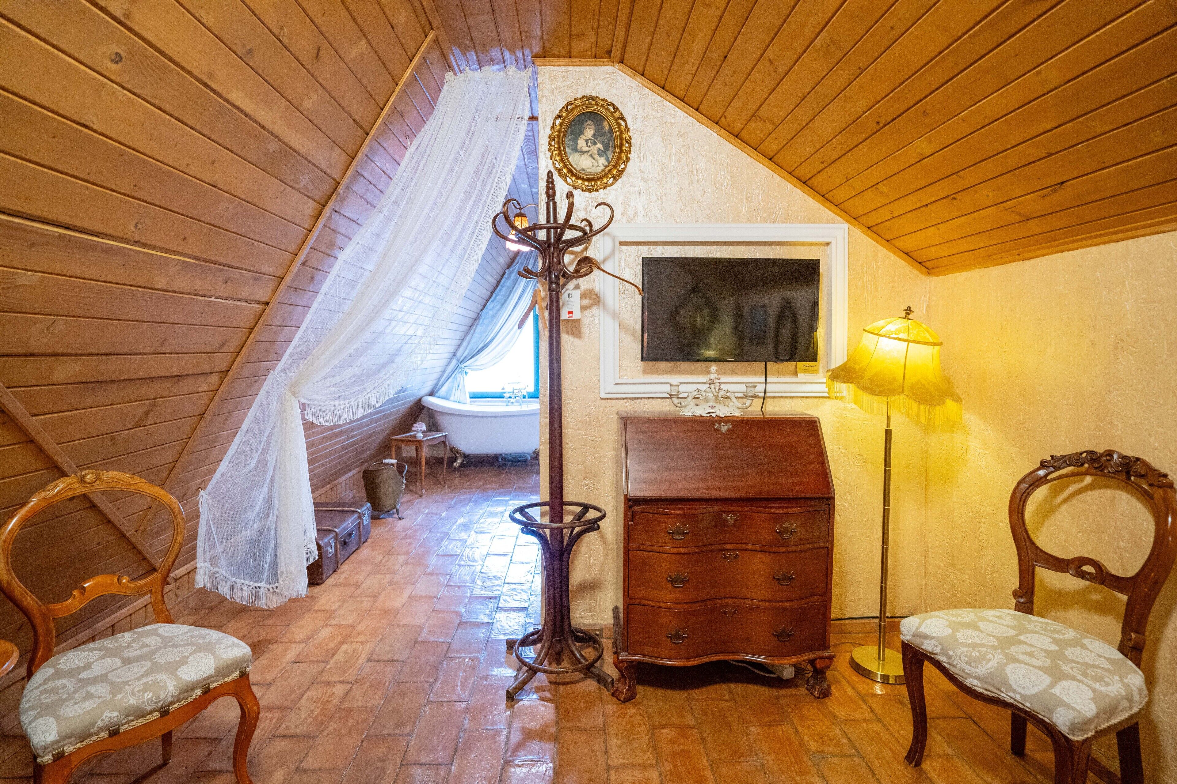 Un dormitorio acogedor en el ático con vigas de madera, una cama, un aparador, dos sillas, una lámpara de pie y una ventana con vistas al exterior.