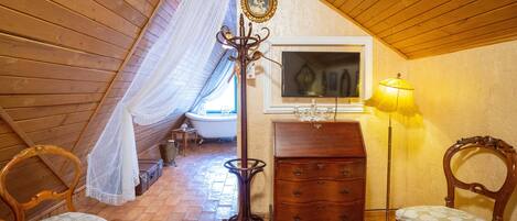 Un dormitorio acogedor en el ático con vigas de madera, una cama, un aparador, dos sillas, una lámpara de pie y una ventana con vistas al exterior.