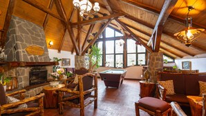 Interior rústico de una cabaña con chimenea de piedra, muebles de madera, mesa de billar y ventanas amplias.