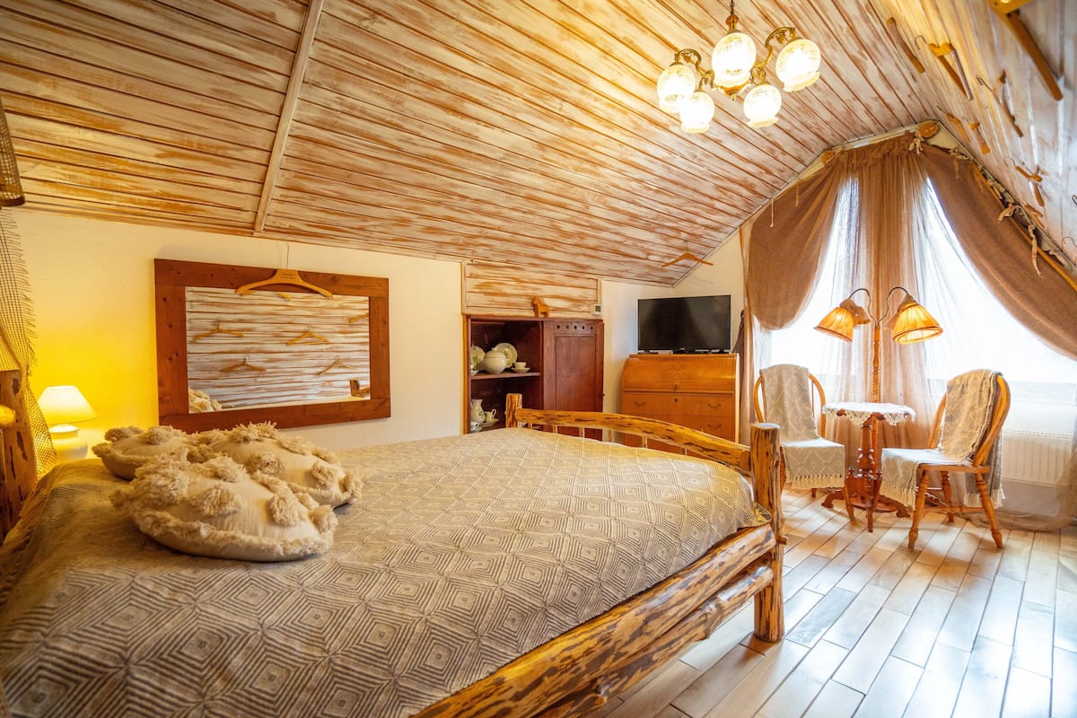 A cozy room with a wooden ceiling, a bed with patterned bedding, a television, and a small table with chairs.
