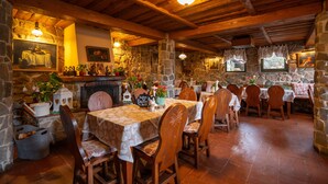 Un comedor rústico con paredes de piedra, vigas de madera y una chimenea. Las mesas están dispuestas con sillas, y hay diversas decoraciones y plantas.
