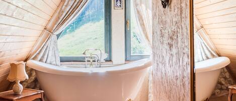 A bathroom with a freestanding bathtub, a small table with a lamp, and a window with curtains.