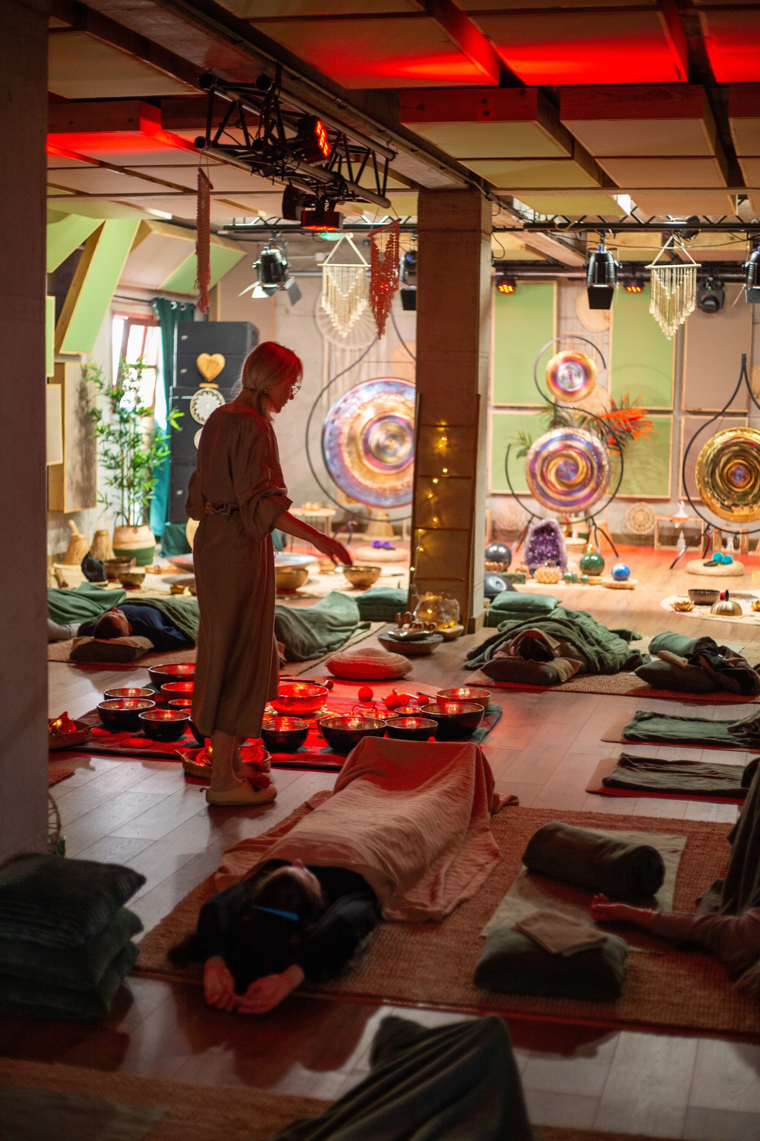 A room with a person standing near a table with bowls and candles, surrounded by mats and cushions on the floor.