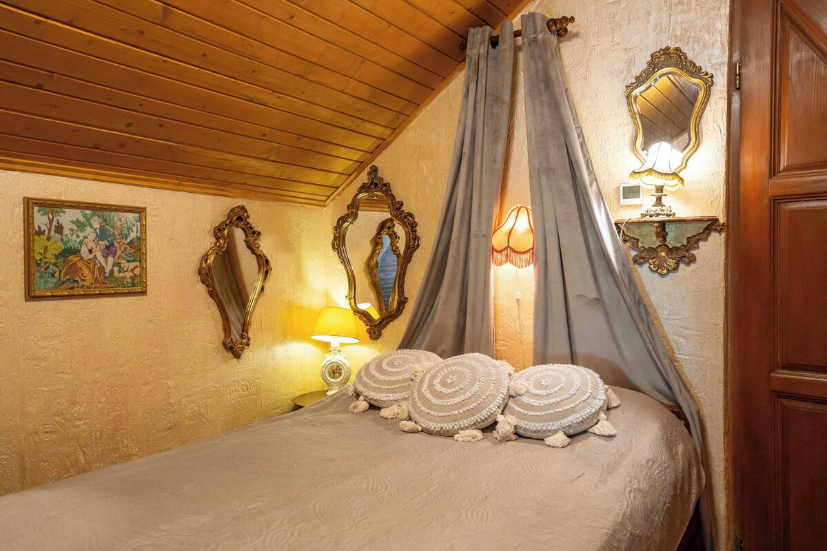 A bedroom with a wooden ceiling, a bed with beige bedding and decorative pillows, a wall-mounted mirror, and a bedside lamp.