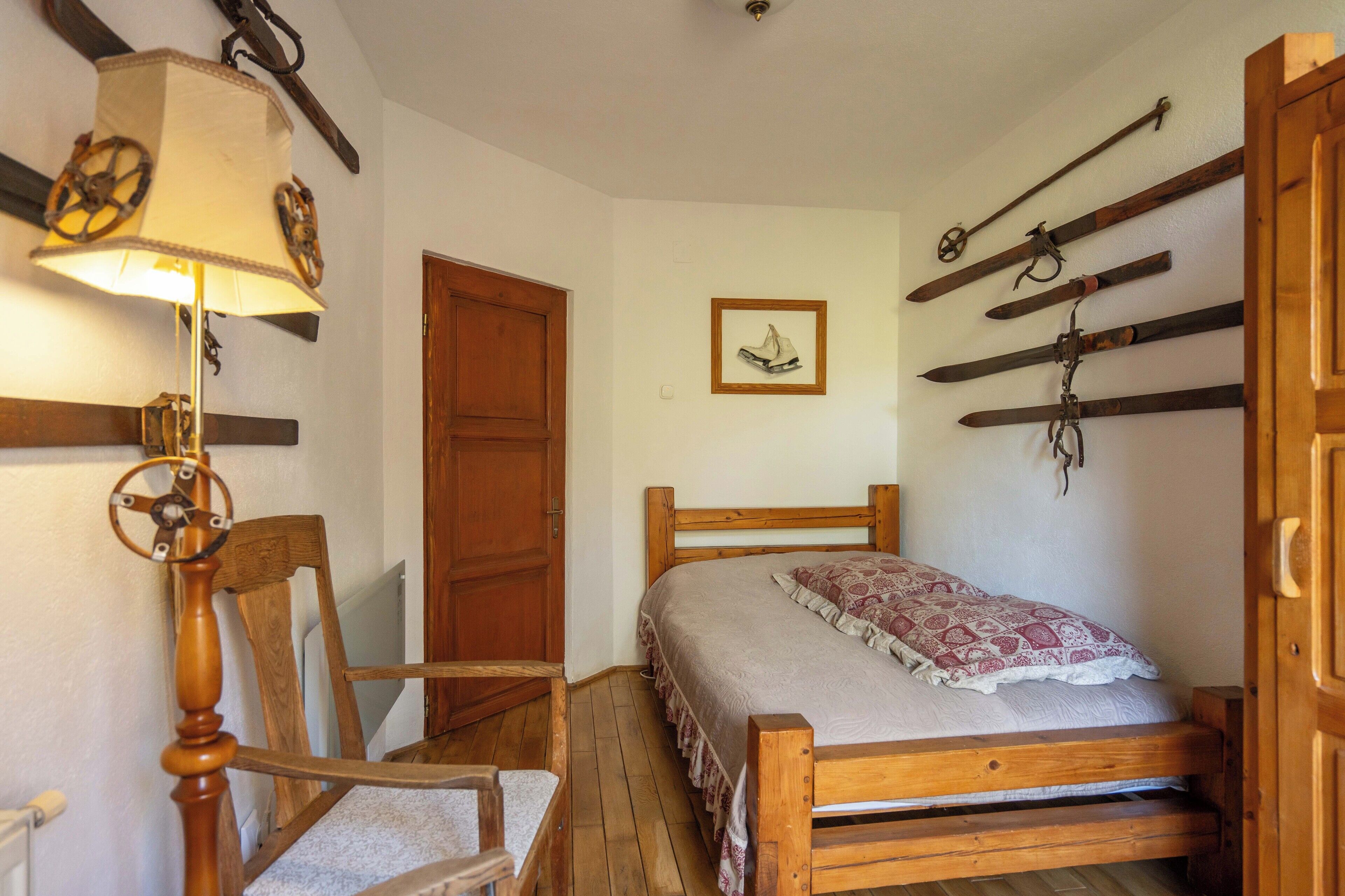 Ein hölzernes Bett mit einem gemusterten Überwurf, ein Holzstuhl, ein Schreibtisch aus Holz und eine Wandlampe.