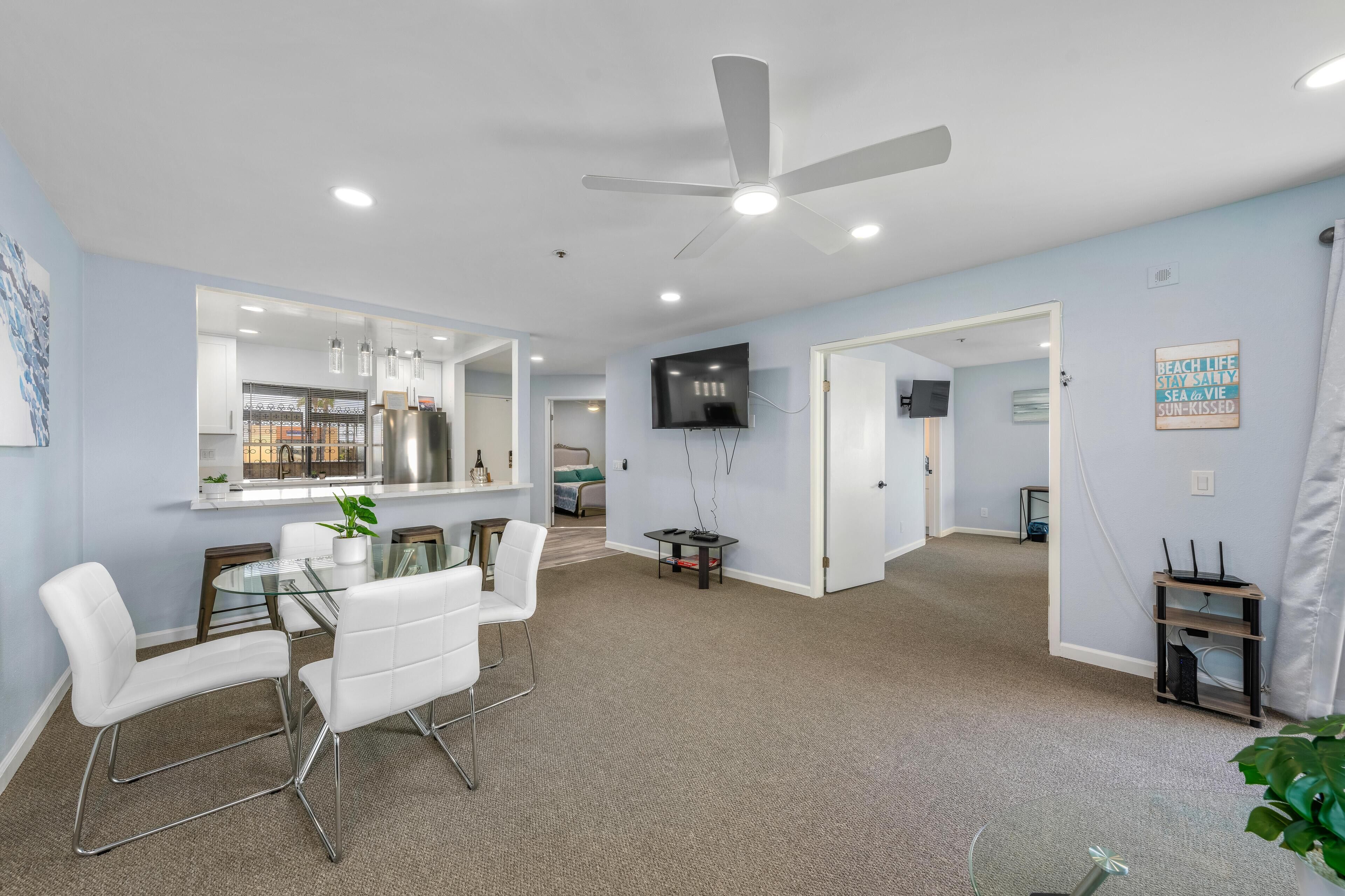 A modern living room with a glass dining table, white chairs, a ceiling fan, and a flat-screen TV mounted on the wall.