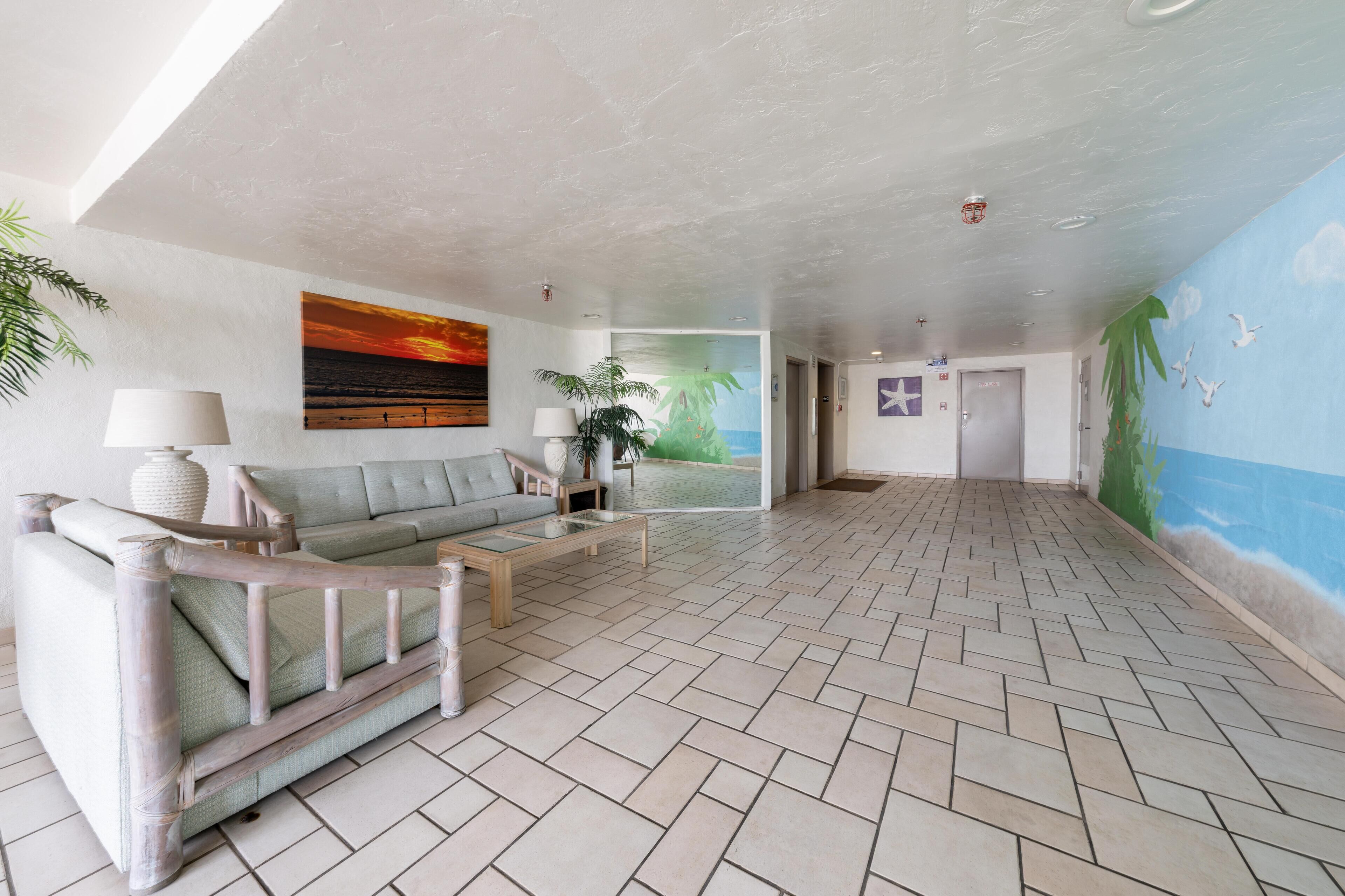 Een ruime lobby met een visgraatvloer, een grijze bank, een houten salontafel en een muurschildering van een zonsondergang.