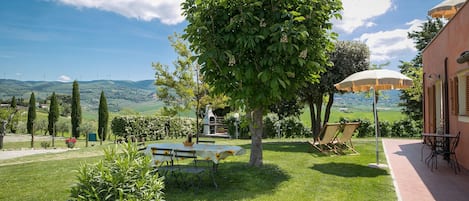Un jardin avec une table et des chaises, un arbre et un bâtiment d’où l’on peut admirer les collines.