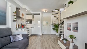 Un espacio de estar moderno con un sofá gris, una cocina con armarios blancos, una escalera y un comedor.