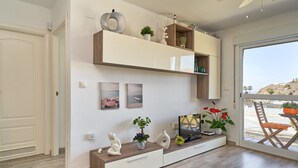 Una sala de estar moderna con televisor, estantes de madera, objetos decorativos y vistas a través de un ventanal.