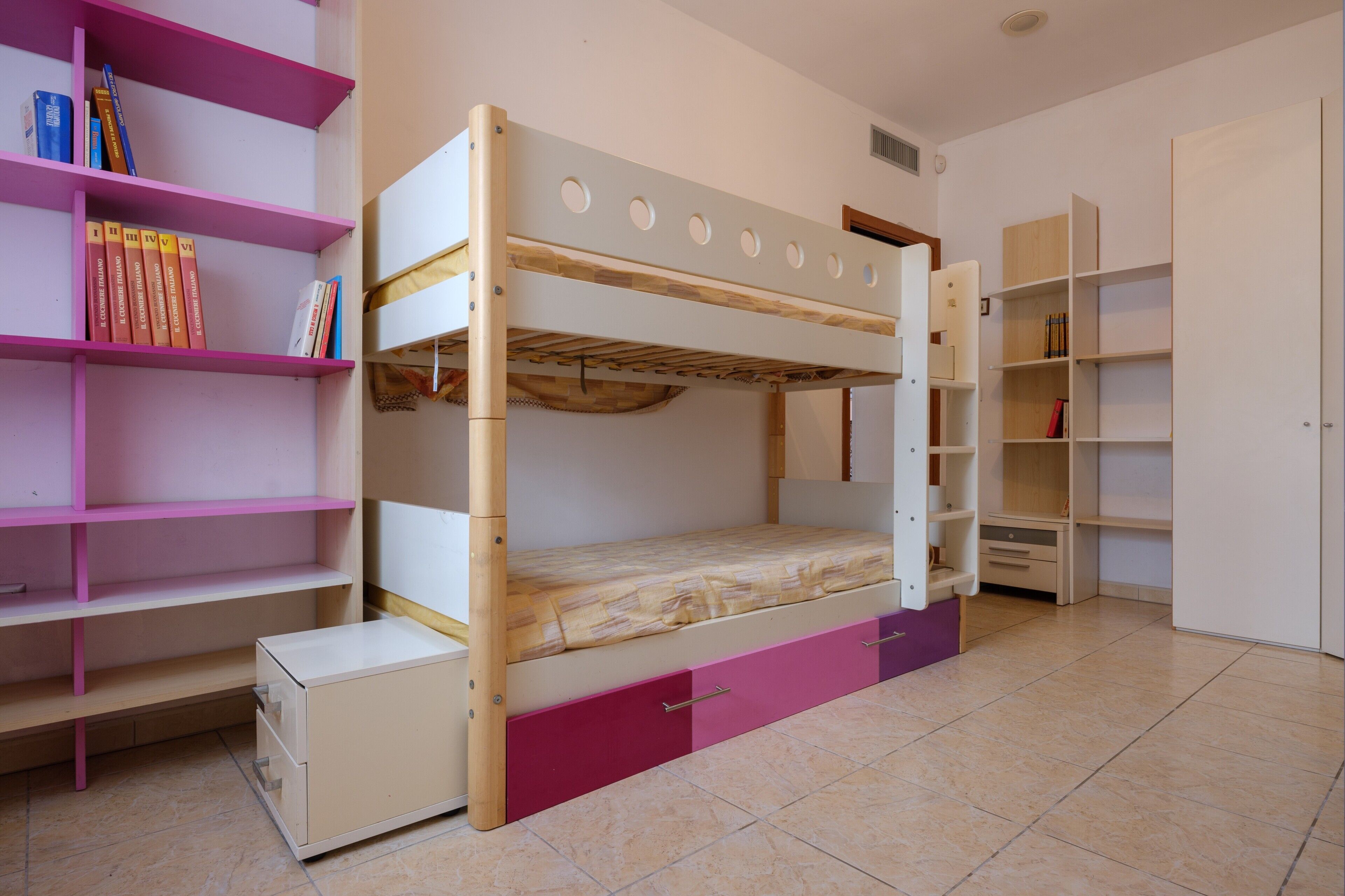 Ein Stockbett mit Holzrahmen und einer Matratze, ein pinkes Bücherregal und ein weißer Schrank mit Schubladen.
