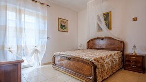 Una camera da letto con un letto in legno, un comodino, una scrivania, una sedia e una finestra con tende leggere.