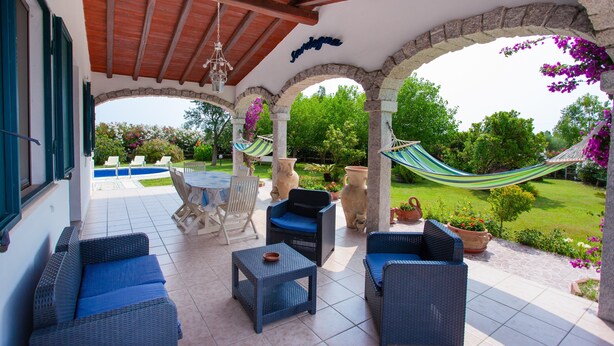 Um alpendre coberto com móveis de vime, uma rede e vista para um jardim e piscina.