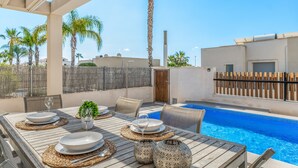 An outdoor dining area with a pool, palm trees, and a modern building in the background. - Villa "Casa Imjo Groundfloor" with Private Pool, Private Terraces & Air Conditioning (Orihuela Costa)