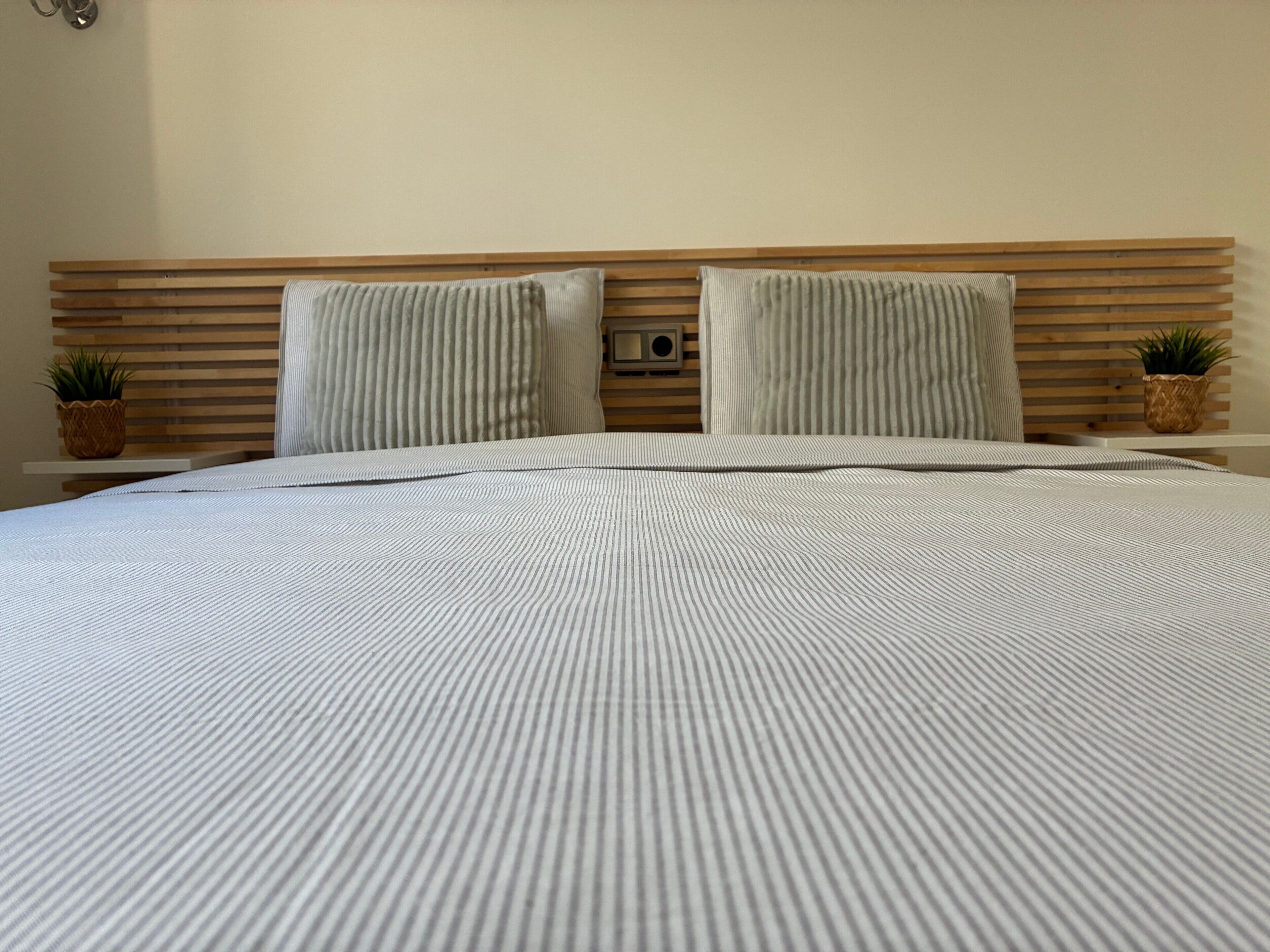 Una cama con colcha a rayas, dos almohadas, cabecera de madera y dos mesitas de noche con plantas en macetas.