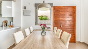 A dining area with a wooden table, white chairs, a vase with flowers, a pendant light, a window with plants, and a wooden door. - Villa "Casa Imjo Groundfloor" with Private Pool, Private Terraces & Air Conditioning (Orihuela Costa)