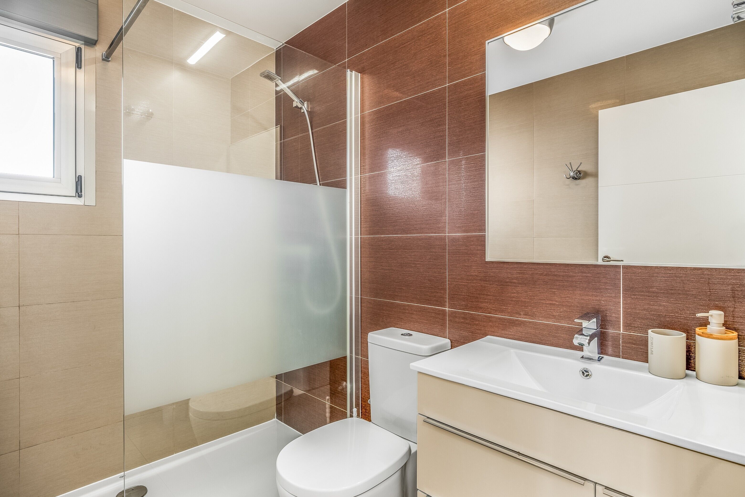 Un baño moderno con cabina de ducha de vidrio, inodoro blanco, lavamanos con espejo y mueble beige.