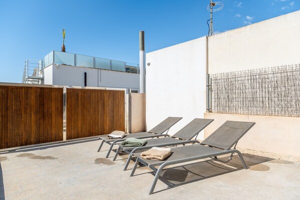A rooftop with four sun loungers, a wooden fence, a white wall, and a building with a glass balcony.