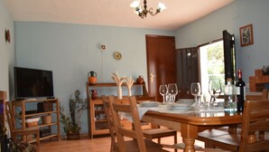 Un comedor con una mesa de madera dispuesta para cuatro personas, rodeada de sillas de madera. A la izquierda, un televisor sobre un mueble bajo, y una puerta que da al exterior.