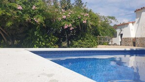 Una piscina con borde de azulejos, rodeada de abundante vegetación y un edificio con techo de tejas.