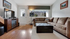 A living room with a wooden floor, a sofa, a television, and a window with a view. - Brookside II B-304 - Sunday River Slopeside Condominium (Newry)