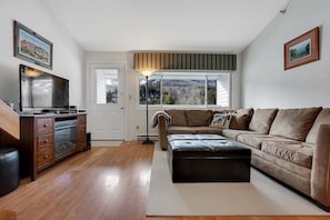 Una sala de estar con piso de madera, un sofá, un televisor y un ventanal con vistas.