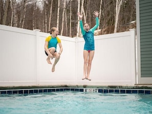 Dos personas saltan a una piscina con una valla blanca y árboles al fondo.