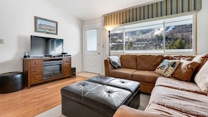 A living room with a TV, sofa, ottoman, and a window with a view of a town. - Brookside II B-304 - Sunday River Slopeside Condominium (Newry)