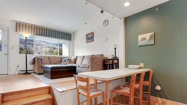 A living room with a sofa, a dining table with chairs, a lamp, and a view through a window. - Brookside II B-304 - Sunday River Slopeside Condominium (Newry)