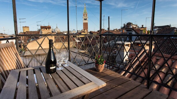 Une table en bois, une bouteille de vin, des verres et une plante en pot sont posés sur un balcon doté d’une rambarde en métal.