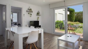 A modern, well-lit room with a white dining table, chairs, and a small white table. There is a TV on a stand, a plant on the wall, and a view of a garden through a sliding glass door. - Apartment with pool and parking. Ground floor with garden (Palafrugell)