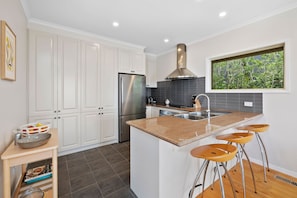 A modern kitchen with white cabinets, a stainless steel refrigerator, a large island with bar stools, and a window with a view of greenery.