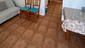 Ein Zimmer mit einer Couch, zwei Holzstühlen, einem Tisch mit gemustertem Tuch und einer Holztür im Hintergrund.