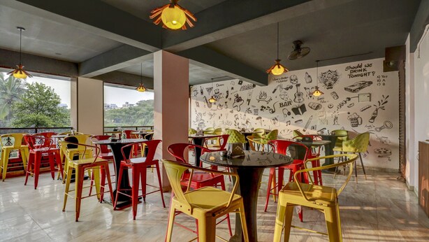 A restaurant with colorful chairs and tables, a mural with various doodles, and large windows offering a view of trees. - Treebo Three Street Inn (Bengaluru)