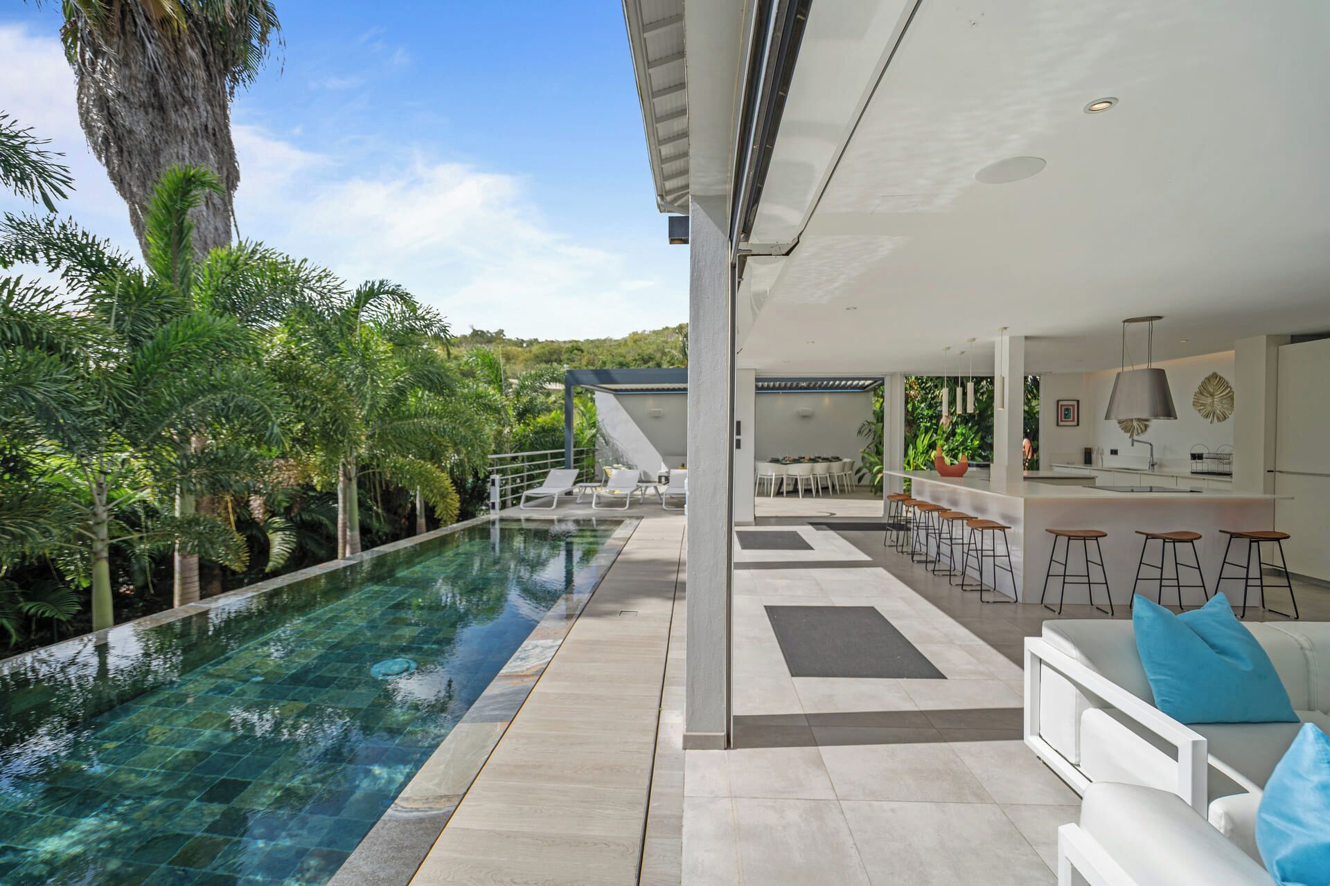 Un espace piscine extérieur moderne offrant une vue dégagée sur la piscine, un bar avec des tabourets et un coin salon avec un canapé et des coussins.